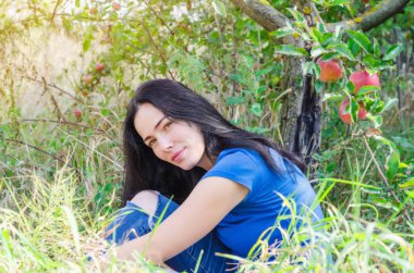 Bahçede bir elma ağacı altında yeşil çimenlerin üzerinde oturan çekici esmer kız. Doğa üzerinde bekletin. Kadın sağlığı. Yaz anıları. Cinsellik, kadınlık