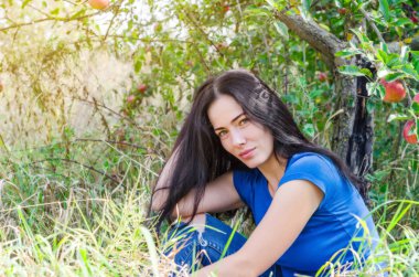 Bahçede bir elma ağacı altında yeşil çimenlerin üzerinde oturan çekici esmer kız. Doğa üzerinde bekletin. Kadın sağlığı. Yaz anıları. Cinsellik, kadınlık