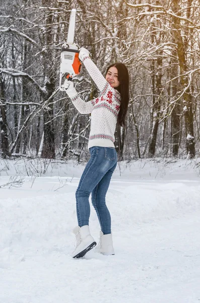 Woman in a sweater and jeans holds a chainsaw in her hands in woods. Deforestation, sawing, lumberjack and forestry equipment.