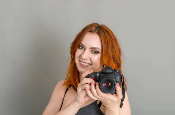 Smiling woman with red hair holds modern camera in her hands. Gray background, creativity, photographer, beautiful