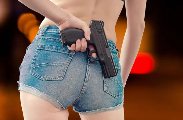 Девушка в джинсовых шортах держит чёрный пистолет за спиной. — стоковое фото