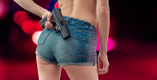 Joven en pantalones cortos de mezclilla está sosteniendo una pistola negra detrás de ella — Foto de Stock