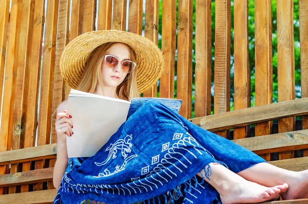 Молодая девушка в солнечных очках в соломенной шляпе читает глянцевый журнал на скамейке на фоне деревянной стены. Летнее настроение, отдых — стоковое фото