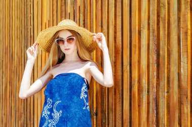 Tahta bir duvarın arka planında hasır şapkalı güneş gözlüklükgenç bir kız. Yaz havası, rahatlama
