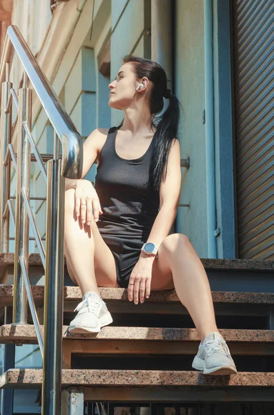 Fitness girl in shorts and T-shirt sitting on stairs with shiny railings. Sports, running, fitness, urban sports.