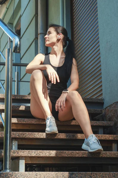 Fitness girl in shorts and T-shirt sitting on stairs with shiny railings. Sports, running, fitness, urban sports.