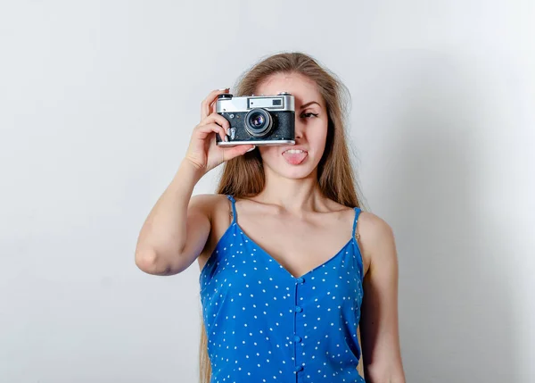 Young beautiful woman is holding an vintage camera and showing the tip of her tongue. She\'s taking a picture wearing a blue summer dress