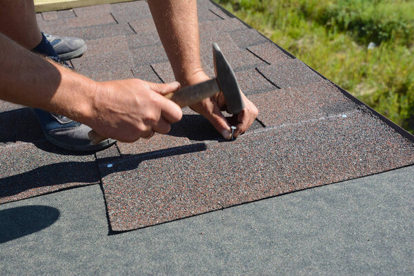 Roofer installing Asphalt Shingles  on house construction roof corner with hammer and nails. Roofing construction with Asphalt Shingles. 