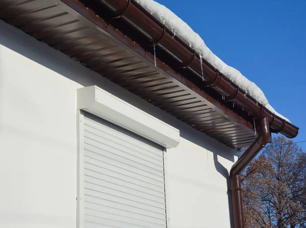 Rain gutter full of ice in winter. Frozen ice in house roof gutter with white rolling shutters for home protection