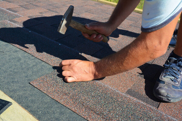 Roofing contractor installing roof tiles, asphalt shingles with hammer and nails. Roofing construction.
