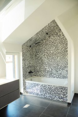 Modern tavan arası tasarımı siyah beyaz camlı mozaik fayanslı duvarlar, siyah mat banyo musluğu ve duş, beyaz küvet ve bir pencere..