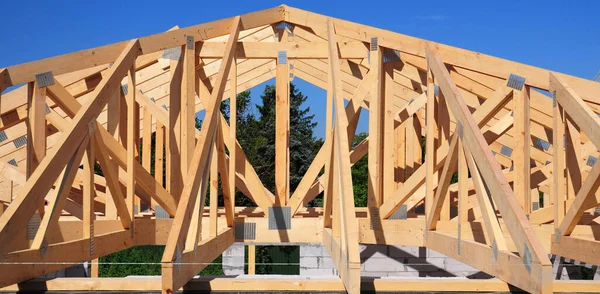 Unfinished roofing wooden frame house construction with roof beams, trusses, timber, braces and eaves of a brick house building against blue sky and forest.