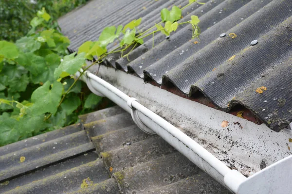 A plastic rain gutter on an asbestos rooftop is cleaned from fallen leaves, dirt and other debris to avoid water overflowing from gutter and protect the house from water damage.