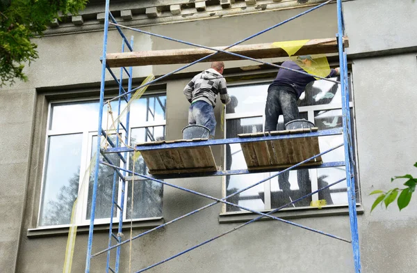 Building contractors on scaffolding are plastering, rendering, applying stucco on the facade of the building with large windows outdoors using reinforcing mesh the prevent cracks.