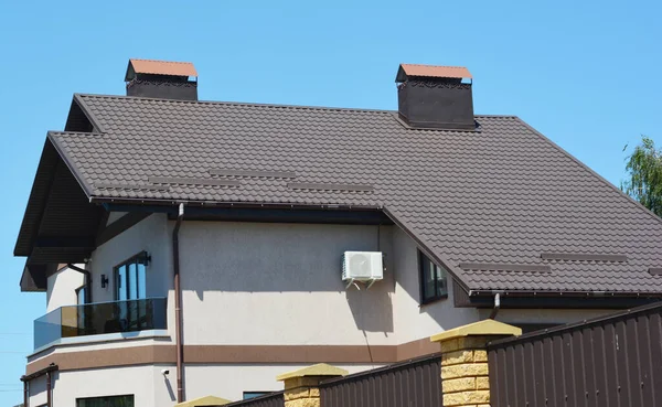 A house with a saltbox, catslide brown metal roof with snow stoppers, chimneys and rain gutter with a downspout, stucco walls with outdoor condenser unit of air conditioner and open balcony.