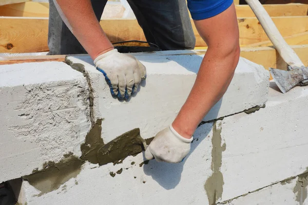 A building contractor is constructing a wall from autoclaved aerated concrete blocks using a trowel and an axe, attaching concrete blocks of home addition to wooden roof beams.