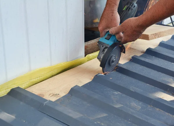 A building contractor is cutting lightweight metal, steel, corrugated, paint coated roofing sheet with an electric angle grinder laying profile sheet on the ground across padded support.