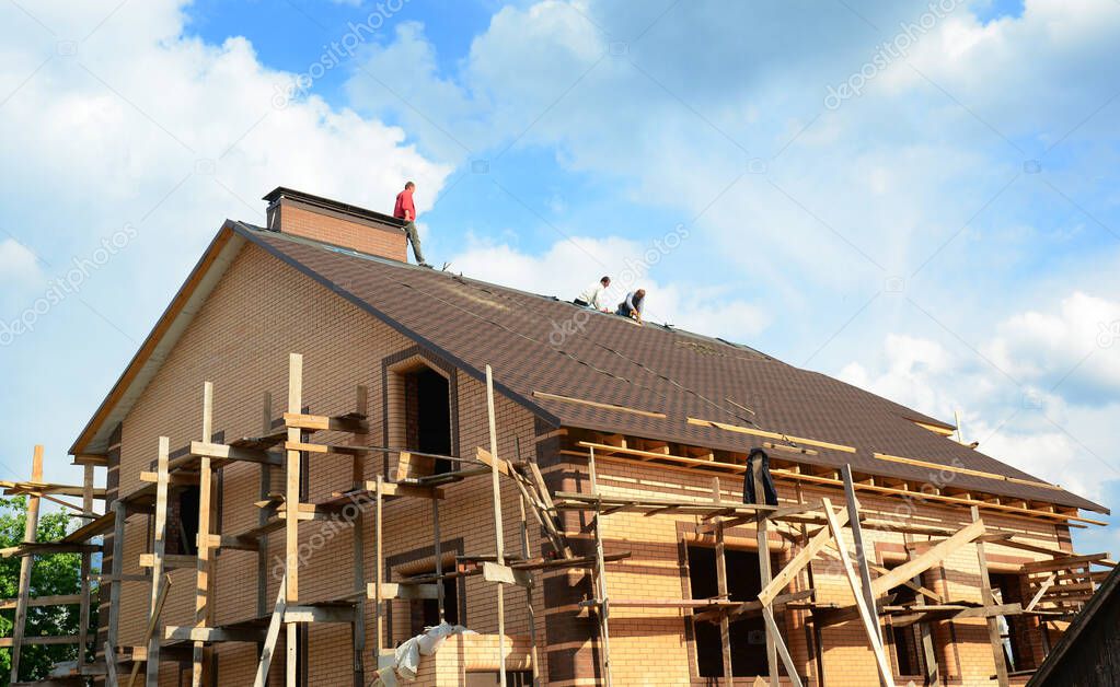 Roofing contractors are finishing installing asphalt roof shingles on the rooftop with a large chimney of a brick house construction with scaffoldings against blue sky.
