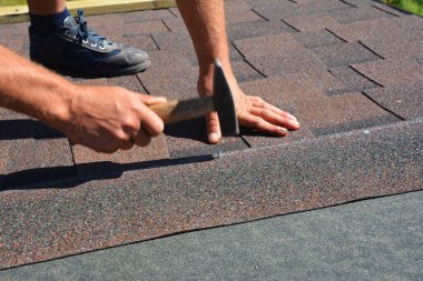 Çatı inşaatı sırasında çatı katında çivi ve çekiçle asfalt kiremitler döşeyen çatı müteahhidi.