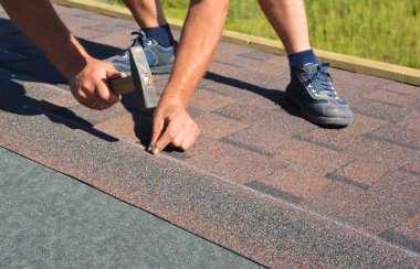 Roofing construction. Roofer contractor nails asphalt shingles on new house rooftop.