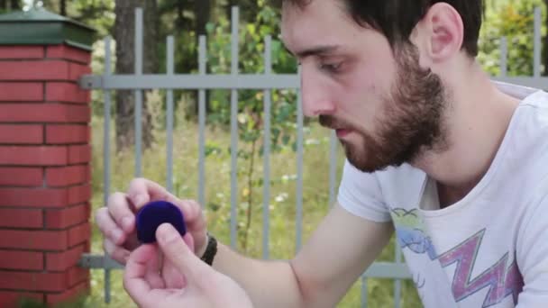 Beau gars ouvre une petite boîte en velours bleu avec une bague de mariage à côté de la clôture — Video