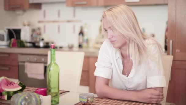 Atraktivní usměvavá blondýna oblečená v bílé košili sedí u stolu v kuchyni — Stock video