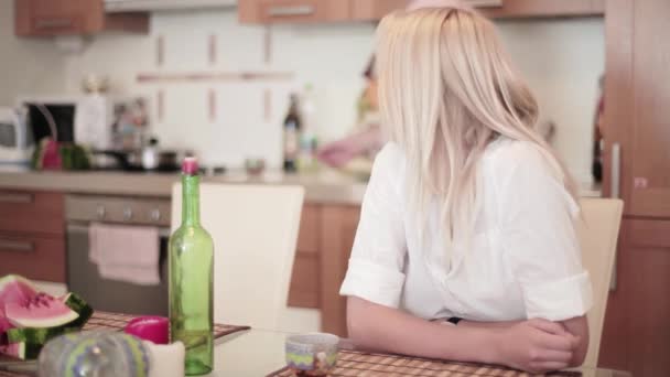 Улыбающаяся блондинка сидит за столом на кухне с молодым человеком, присоединяющимся к ее компании — стоковое видео