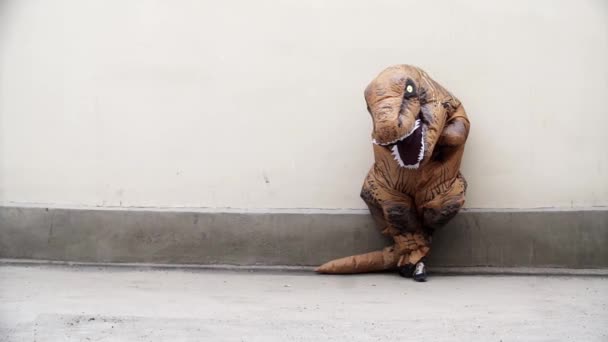Pessoa em t rex mascote traje em pé inclina-se na parede branca — Vídeo de Stock