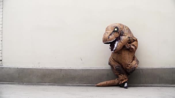 Человек в костюме талисмана динозавра опирается на белую стену — стоковое видео