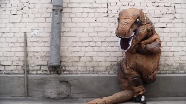 Человек в костюме динозавра стоит, опираясь на стену из белого кирпича — стоковое видео