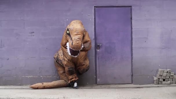 Person im T-Rex-Maskottchen-Kostüm lehnt neben Tür an lila Wand — Stockvideo