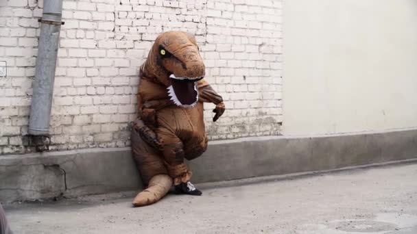 Человек в костюме талисмана динозавра опирается на стену из белого кирпича и убегает. — стоковое видео