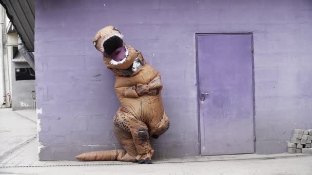 Man in dino mascotte kostuum permanent leunt naast deur op paarse muur — Stockvideo