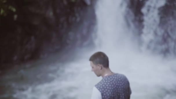 Back view of young man with cigarette standing in front of small waterfall. — Stock Video
