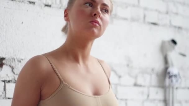 Cute blonde girl dressed in beige leotard stands by barre on white brick wall. — Stock Video