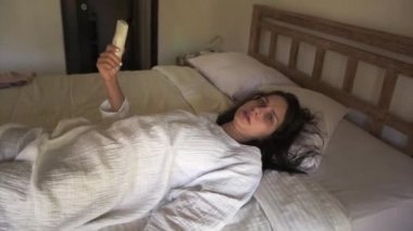 Smartphone elinde yatakta yatıyor tutan beyaz genç ve güzel kadın beyaz elbiseli koyu ıslak saçlı konuşuyor ve gün ışığı yatak odasında kamera bakar.