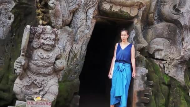 Cute woman in blue clothes stands next to cave with carved face-shaped entrance — Stock Video