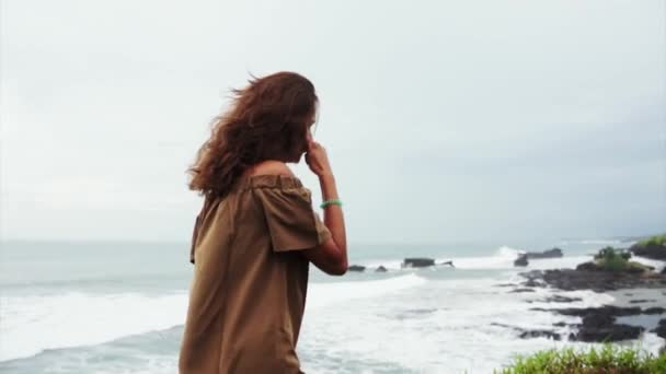 Mujer atractiva con el pelo castaño rizado se encuentra en el acantilado y mira las olas del mar — Vídeo de stock