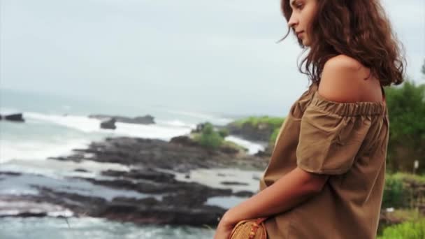 Mujer madura linda con pelo rizado marrón se encuentra en el acantilado y mira las olas del mar — Vídeo de stock