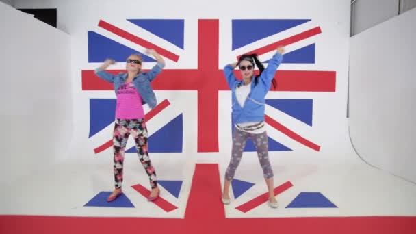 两个迷人的微笑的女孩 长着黑色和红色的头发戴墨镜 穿着明亮的衣服是同步和动态跳舞在英国国旗的背景下 — 图库视频影像