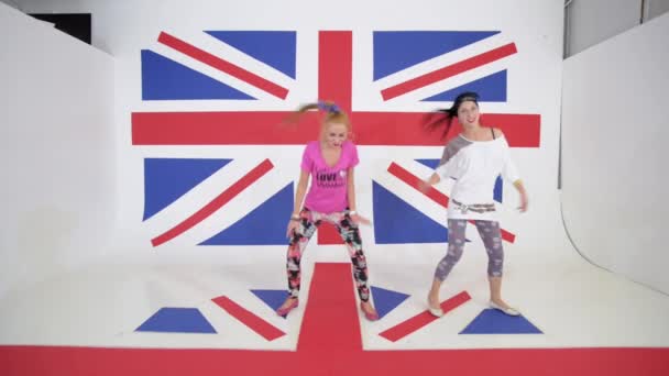 Két vicces aktív lányok dinamikusan táncolnak a háttérben a brit zászló — Stock videók