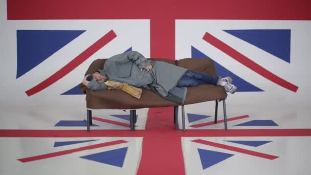 Mann mit Mütze und grauem Mantel liegt auf Sofa und zerkratzt Zimmer mit UK-Fahne. — Stockvideo
