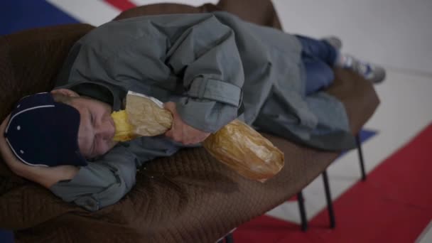 Людина в синій шапку і Сіре пальто лежить на дивані і їсть хліб в кімнаті з Великобританії прапор. — стокове відео