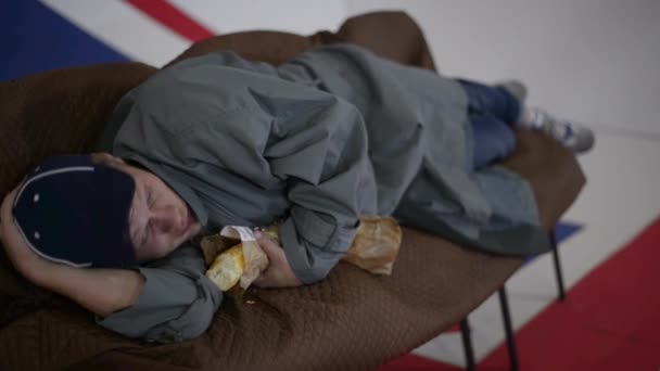 Uomo in berretto blu e cappotto grigio sdraiato sul divano e mangia pane in camera con bandiera britannica . — Video Stock