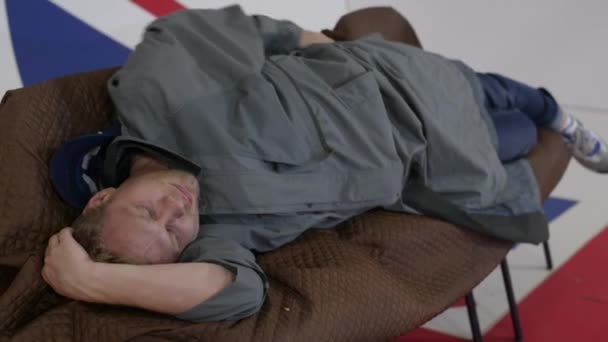 Uomo in lungo cappotto grigio sdraiato sul divano e si gratta la schiena in camera con bandiera britannica . — Video Stock