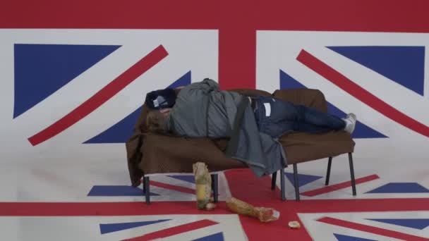 Man in cap and grey coat lies on sofa and turn around in room with UK flag. — Stock Video