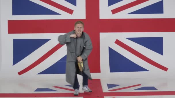 在长大衣的高加索年轻男性持有瓶和舞蹈在房间与英国国旗. — 图库视频影像