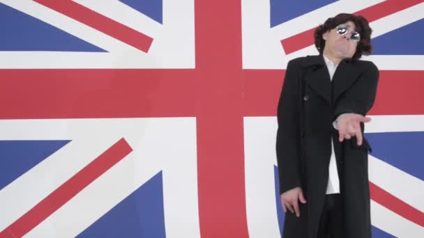 カツラ、サングラス、コートと変な男が英国旗のスタジオのカメラに歌う — ストック動画
