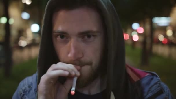 Mladý roztomilý vousatý kavkazské muže v hoodie kouří a dívá se na kameru — Stock video