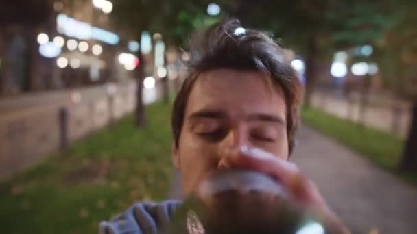 Drunk guy in sweater is drinking from bottle in night street in front of camera. — Wideo stockowe